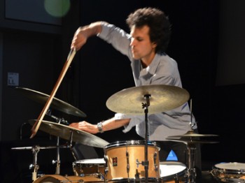 A man plays on a drum kit with a cello bow.
