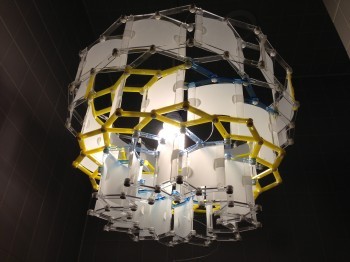 A large lamp made of geometrically interlocking rods and opaque plastic panels.