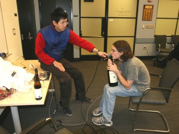 A student holds a microphone up to another student blowing across the top of a wine bottle.