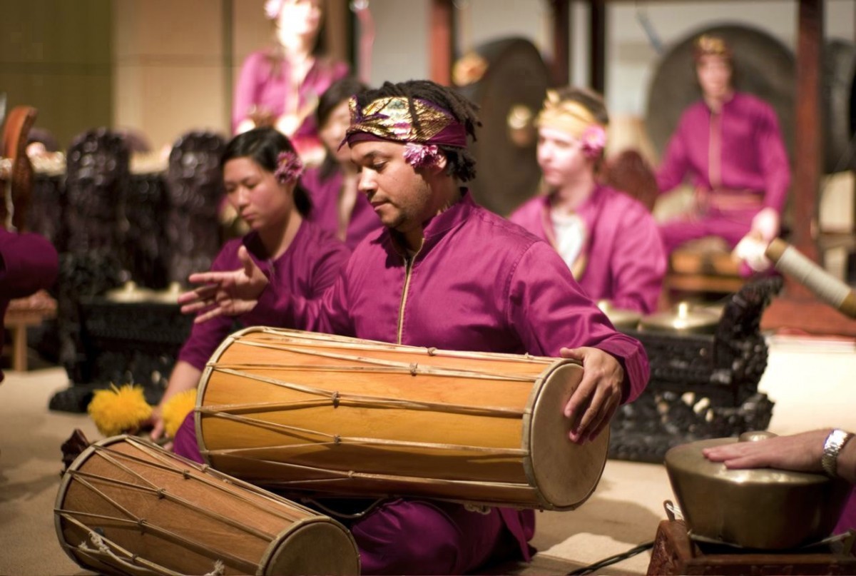 A musician wearing a magenta outfit plays a long drum. Other musicians play instruments in the background.