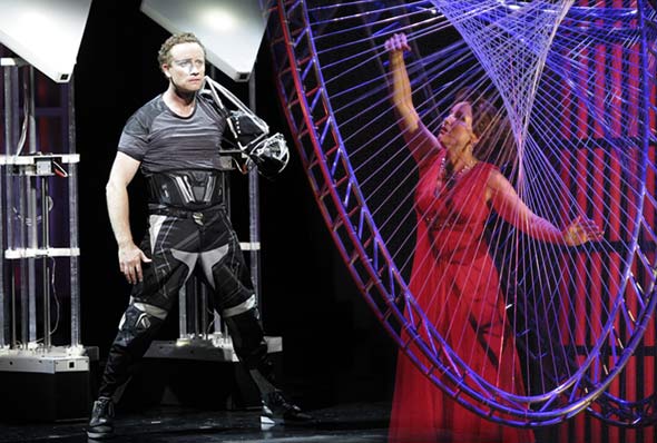 Performers on stage next to a complex metal and wire set piece.