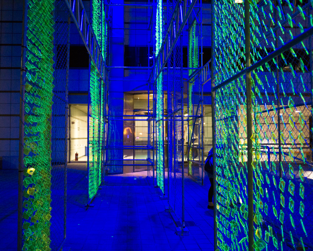 A series of chain metal panels hang vertically outside the MIT Wiesner Building at night, small lime green shapes suspended on the panels appear to glow in the light from the building nearby.