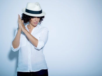 Luciana Souza stands against a pale blue wall while wearing a white panama hat.