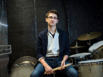 A student holds drumsticks next to a drum kit.