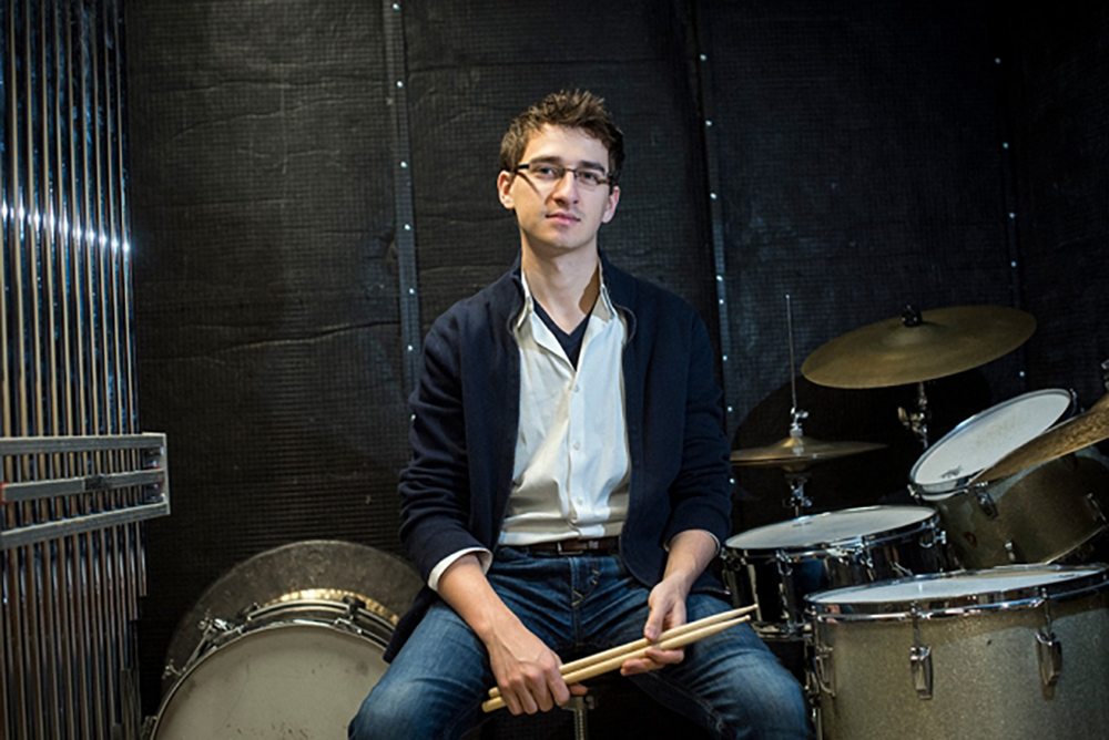 A student holds drumsticks next to a drum kit.
