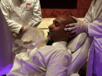 Artists in white lab coats interact with a man in a reclining chair.