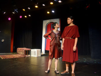 Two actresses in red costumes perform an argument on stage.