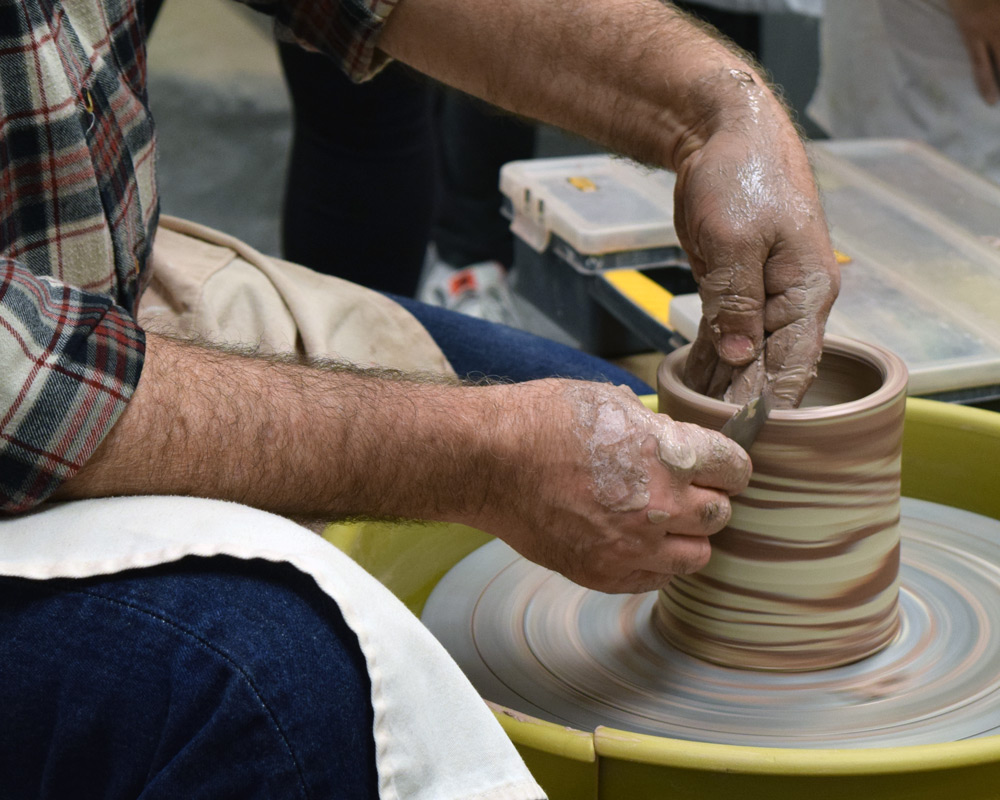 Close up color photograph of two hands using a metal rib to shape a cylinder of brown and white marbled pottery on a potter's wheel.