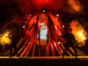Two students in black costumes perform on a stage with red and yellow lights and smoke effects.