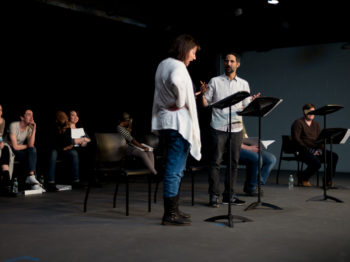 Two actors read from scripts while others look on.