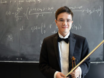 A student holds a violin and bow in front of a chalkboard.