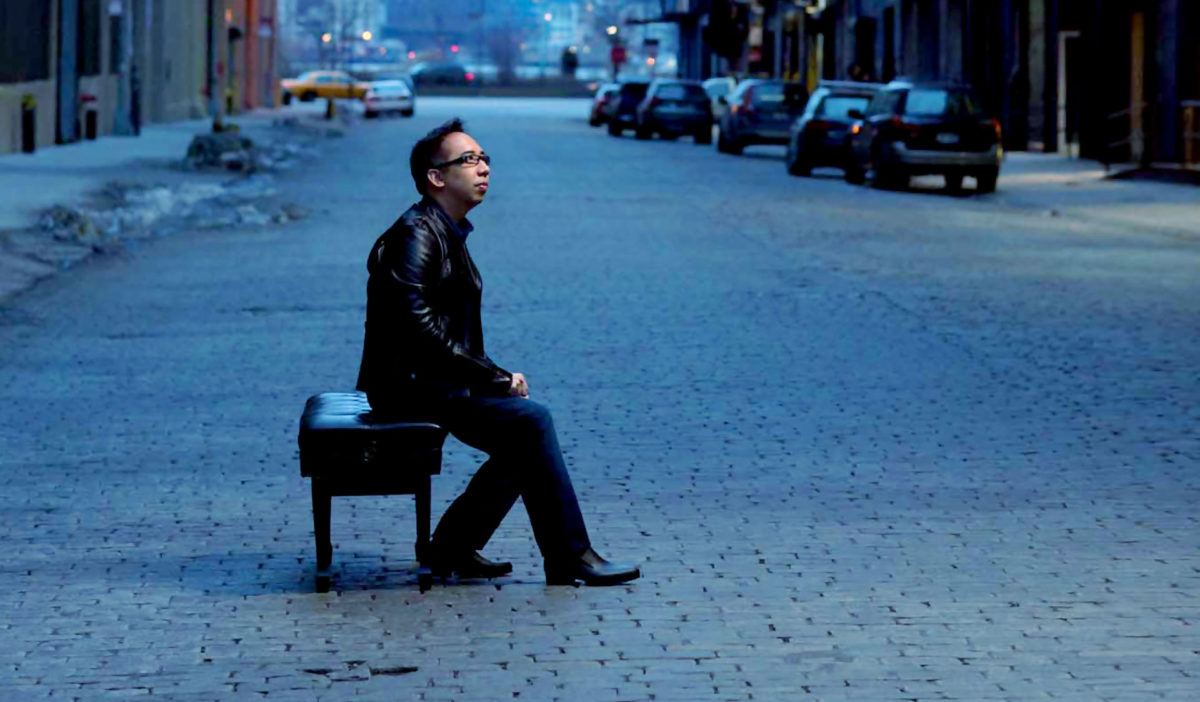 Joel Fan poses on a piano bench on a street.