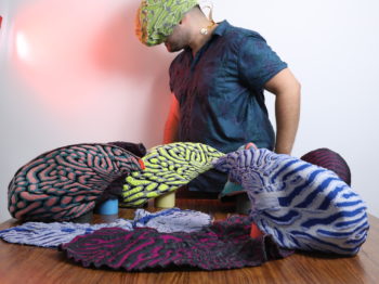 A student wearing a colorful headpiece sits surrounded by other colorful patterned sculptures.