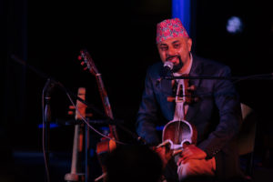 Lochan Rijal speaks into a microphone while sitting on stage and holding a traditional Nepali instrument.