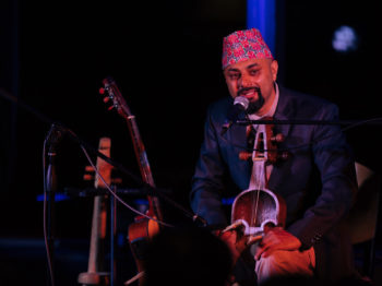 Lochan Rijal speaks into a microphone while sitting on stage and holding a traditional Nepali instrument.