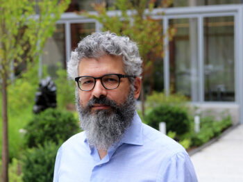 Keeril Makan stands in an MIT courtyard with green foliage in the background and looks directly into the camera.