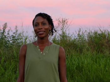 Carla Lyndale Bishop stands in tall grass with a pink sunset lit sky in the background.