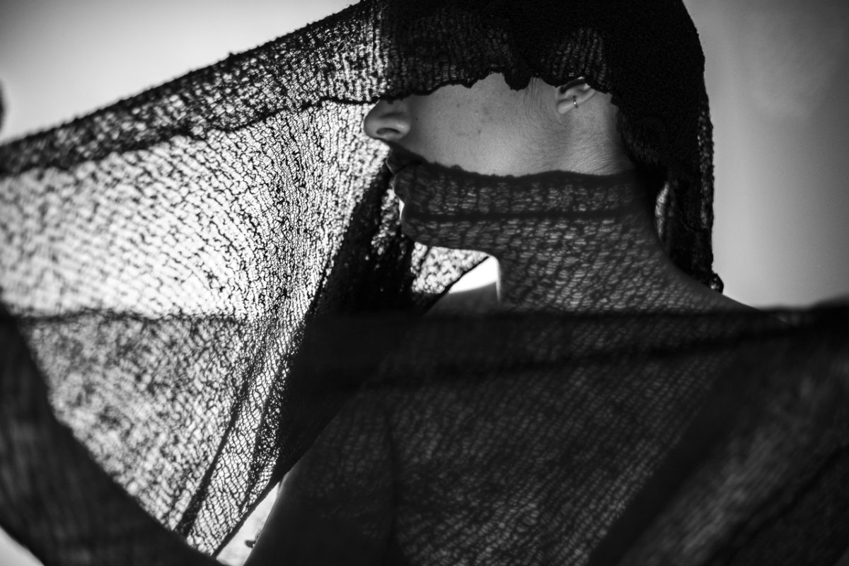 Black and white photograph of a side profile of a figure wearing a translucent black scarf drapped around their head and shoulders.