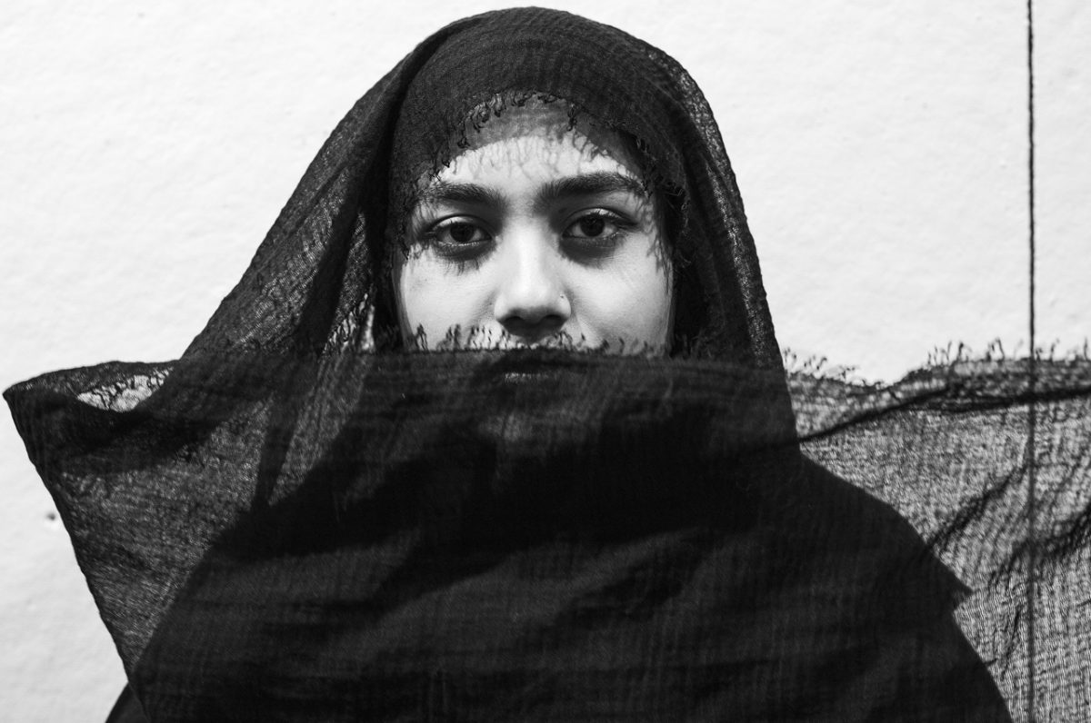 Black and white photograph of a figure facing the camera wearing a translucent black scarf drapped around their head and covering their mouth.