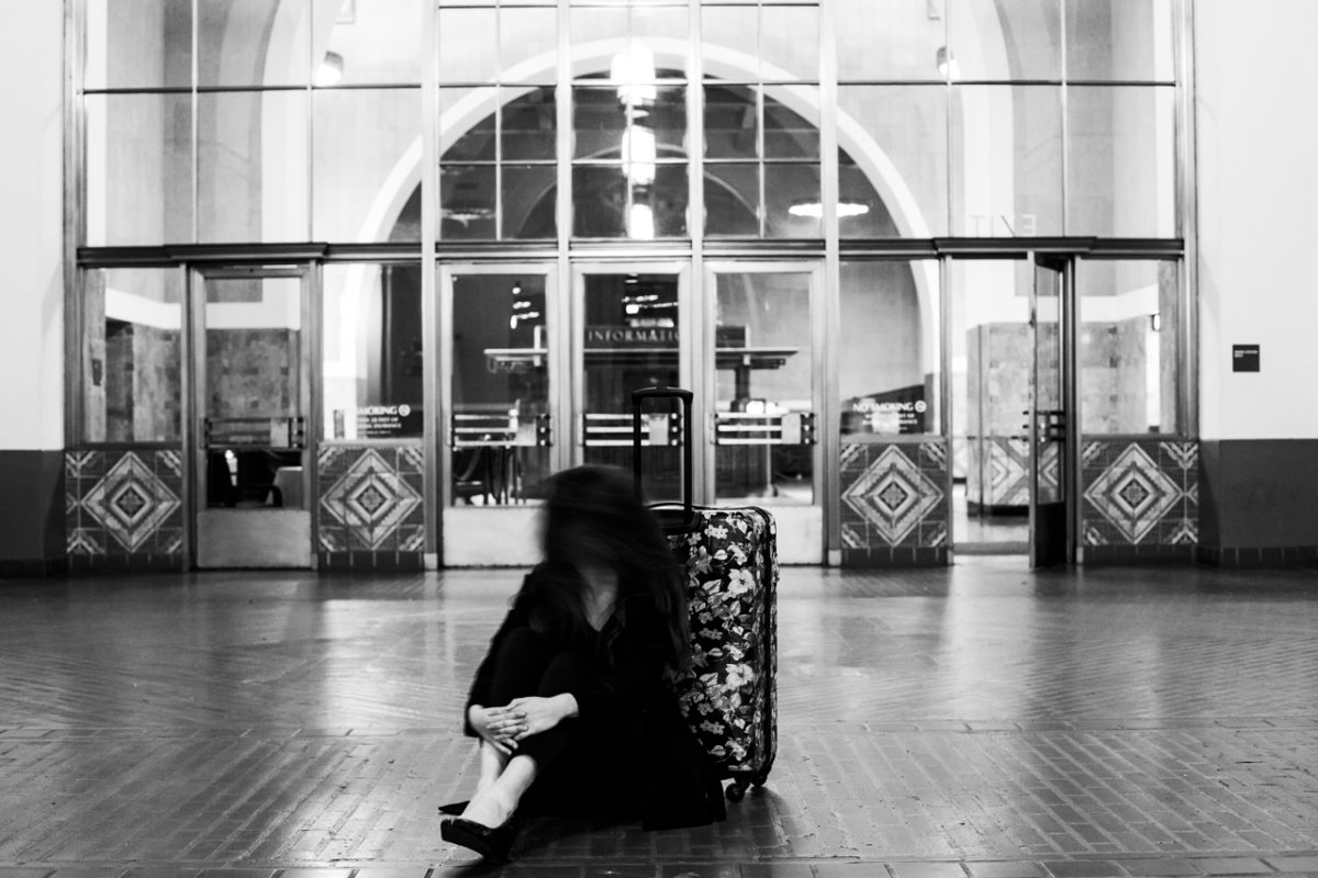Black and white photograph of a figure sitting on the ground in a transit station with both of their hands around their knees and leaning against a floral patterned suitcase.