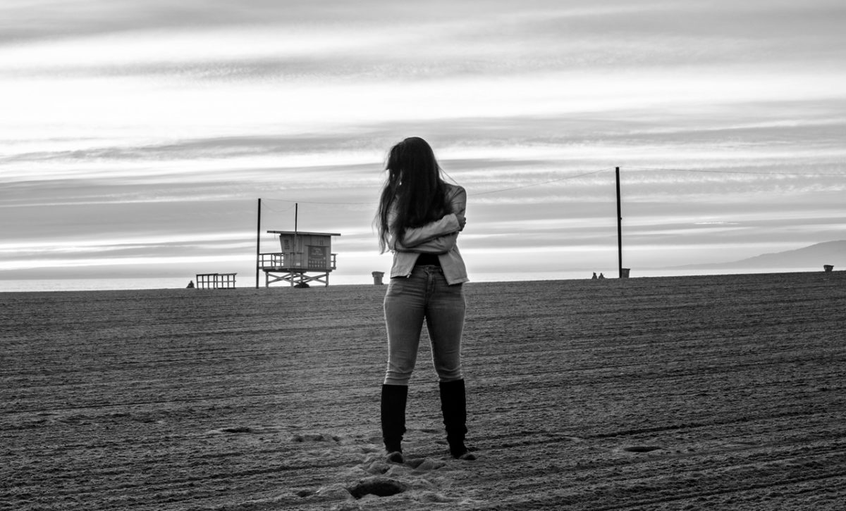 Black and white film photograph of a figure in jeans and tall boots on a beach with a lifeguard tower in the distance.