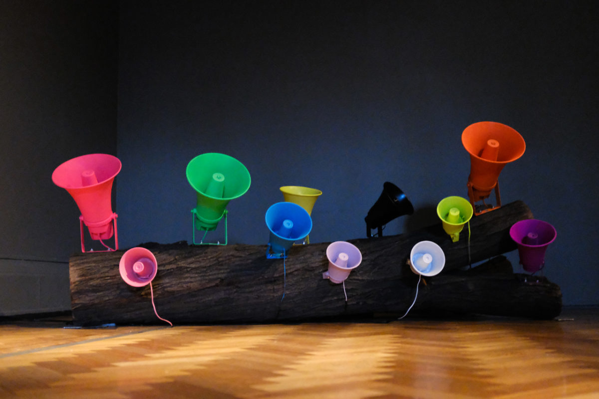Installation view of ten multicolored speakers on a hardwood floor.