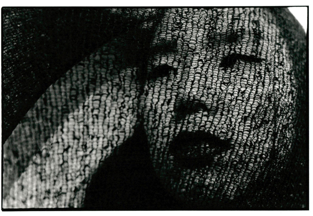 Close up black and white photograph of a young person's face obscured by loosely woven fabric.