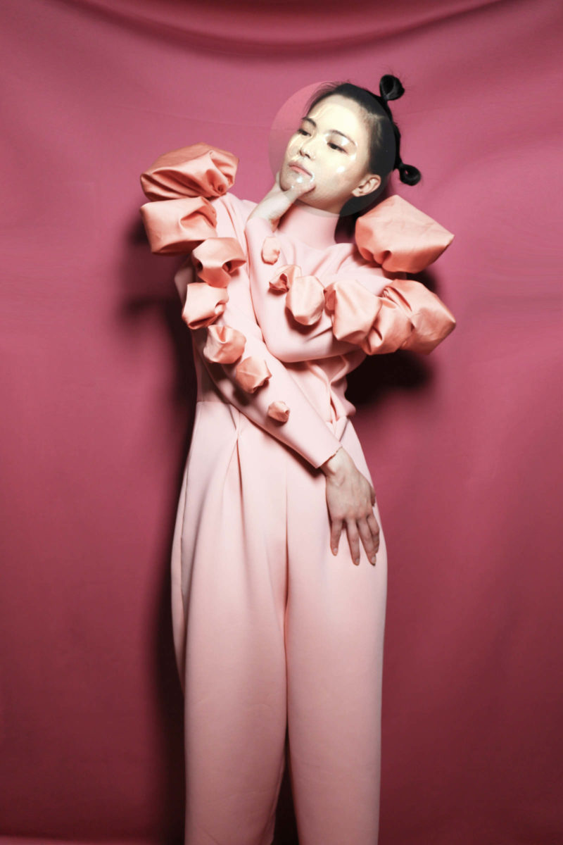 Rae Yuping Hsu wears the pink OOloI spacesuit and stands in front of a dark pink wall.