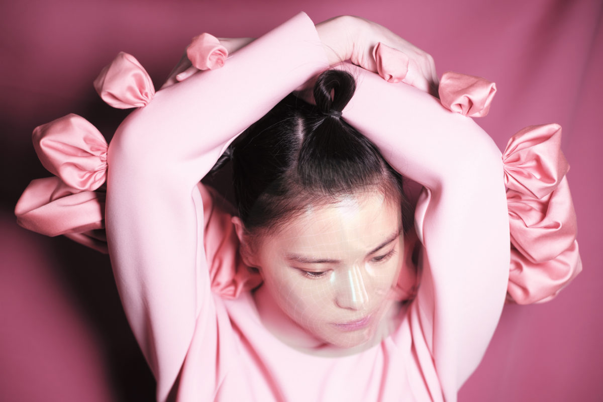 Rae Yuping Hsu wears the pink OOloI spacesuit with arms crossed over their head and stands in front of a dark pink wall.