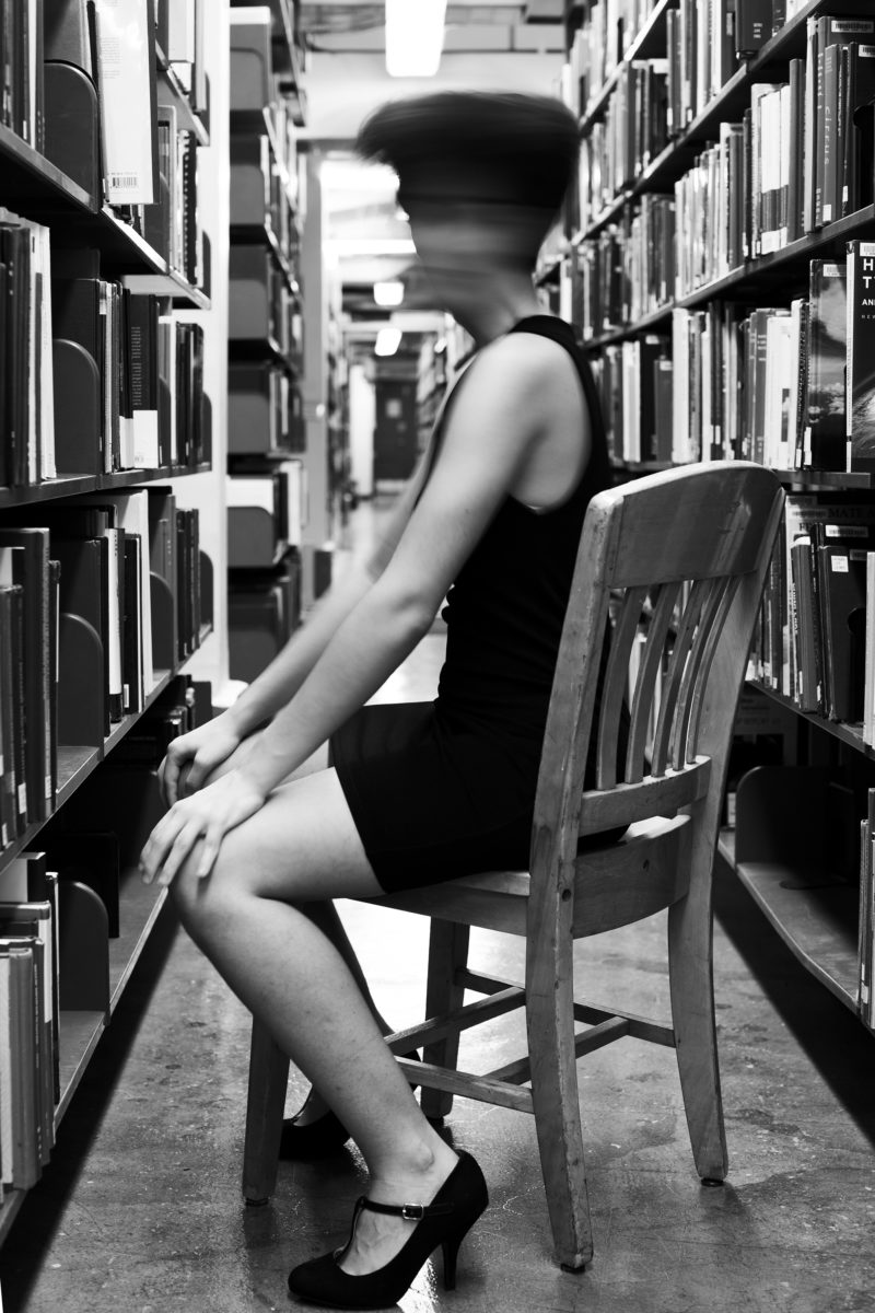 A figure wears high heels and sits in a chair in an aisle of bookcases, their face blurred by movement during a slow camera exposure.
