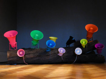 Installation view of kinetic-sonic sculpture, eleven colorful speakers attached to trunk of tree laying on a wood floor.