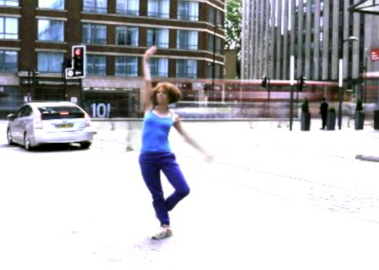 Adesola Akinleye dances on a busy street sidewalk while the background motion of cars and people are blurred.