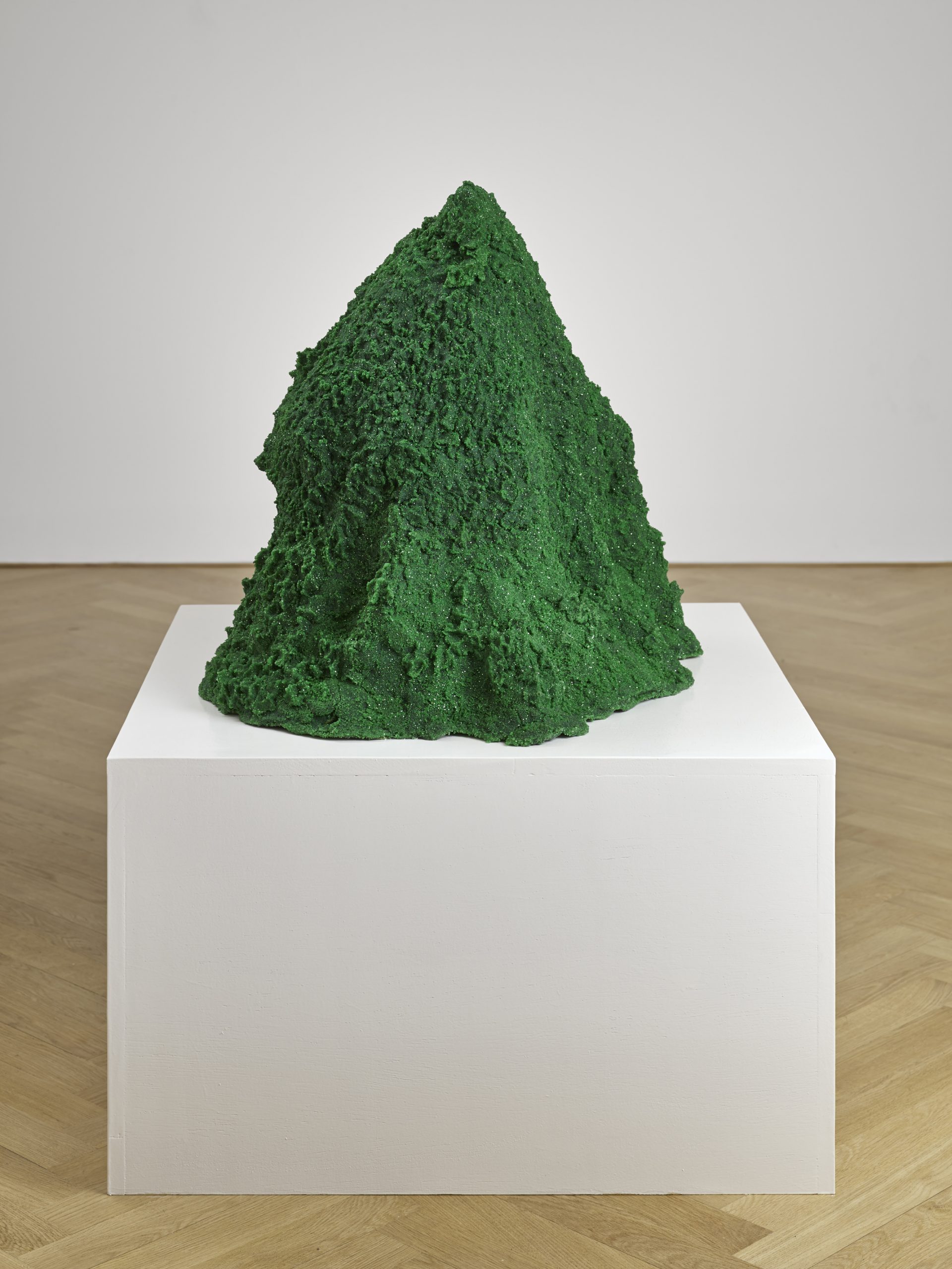 A mountain-like green mound of colored sand, gold, glitter, and crystals sits on a white pedestal in an art gallery.