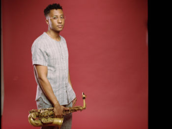 Braxton Cook stands in front of a red background, holding his alto sax down at his waist.