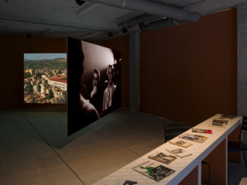 A table with booked laid out on it next to two large photographs of people and cityscapes displayed in an art gallery.