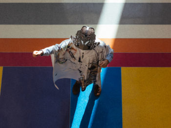 A person wearing a decorative mask and robe standing on a colorful terrazzo floor holds their fist in the air.