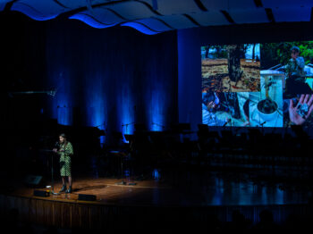 Talia Khan speaks into a microphone on stage in Kresge Auditorium with a large projection of images of Brazil behind her.
