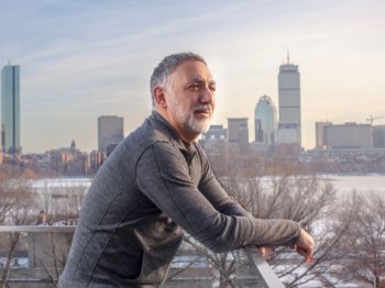 Hashim Sarkis in a gray top at a roof top in Cambridge with a morning view across Charles River view of Boston.
