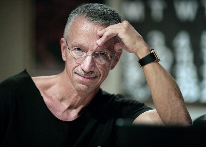 Keith Jarrett in a black shirt with a hand on his forehead smiling into the camera.