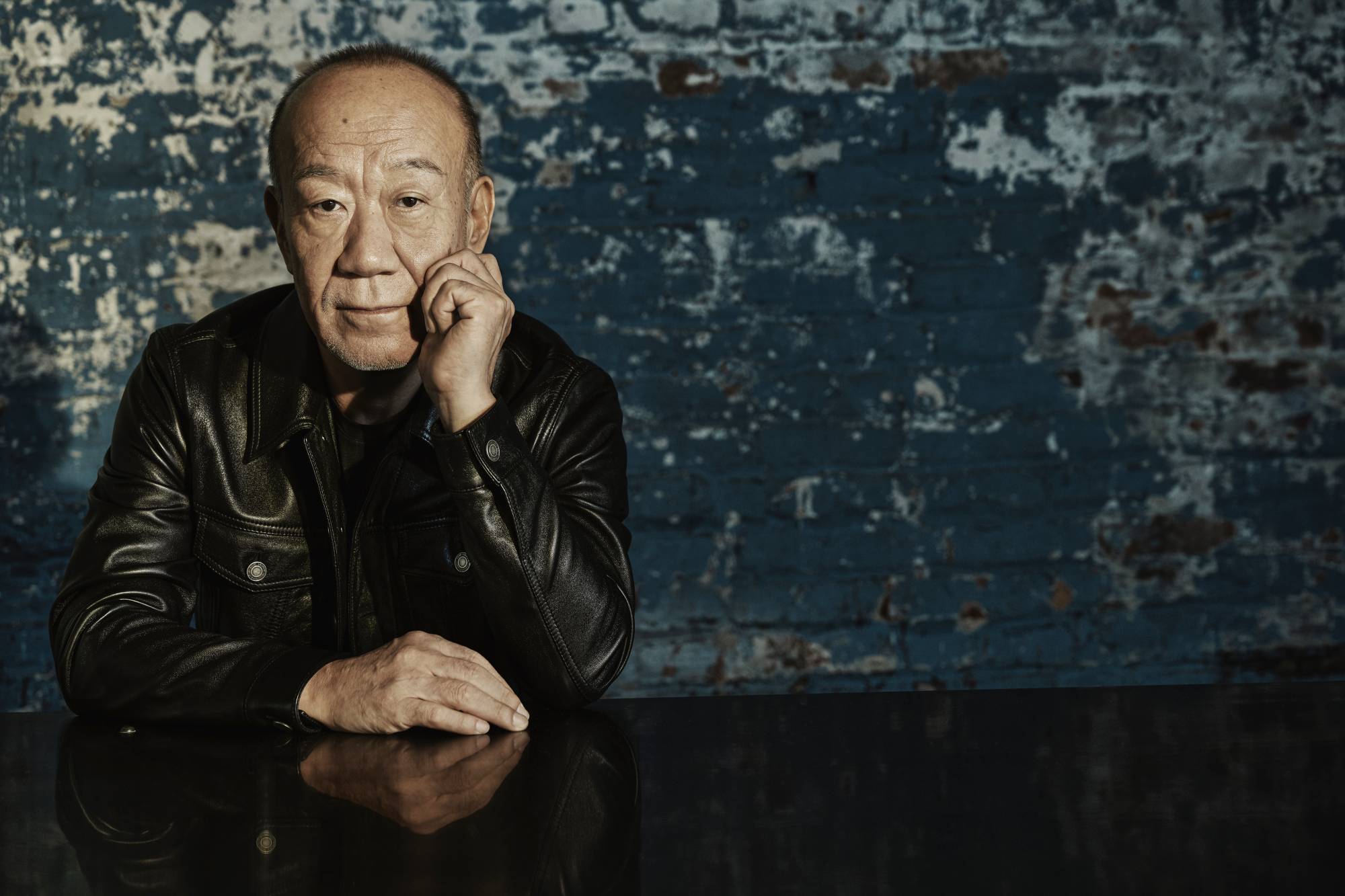 Joe Hisaishi leaning on a piano in front of a blue wall smiling into the camera.
