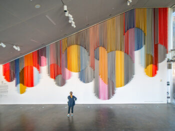 Single gallery visitor stands facing a large colorful installation of circles on a white wall.
