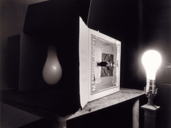 A cardboard camera obscura sits on a table in a darkened room, transferring the image of a lightbulb through a small hole to the interior wall of the box.