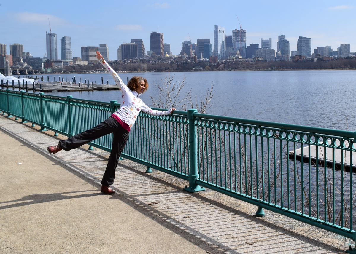 Adesola Akinleye stands in front of the Charles River and Boston Skyline with an arm and a leg raised.