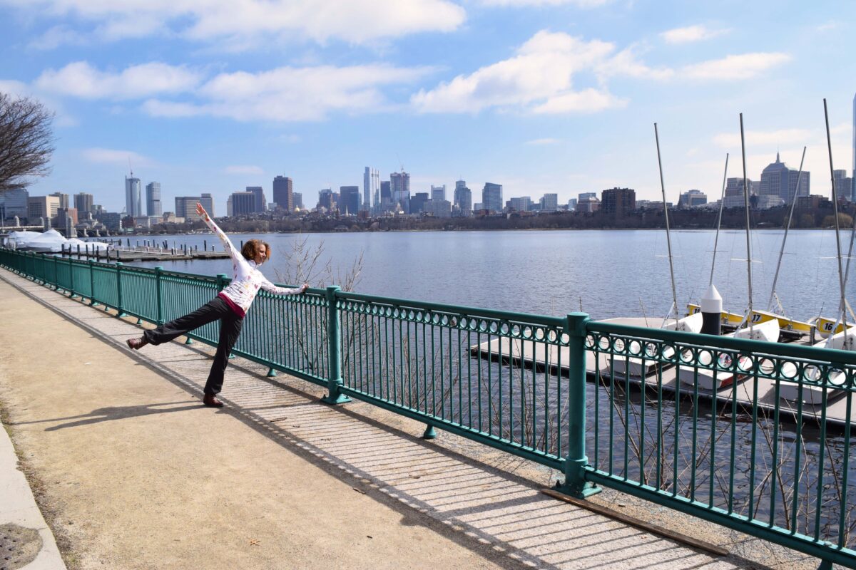 Adesola Akinleye stands in front of the Charles River and Boston Skyline with an arm and a leg raised.