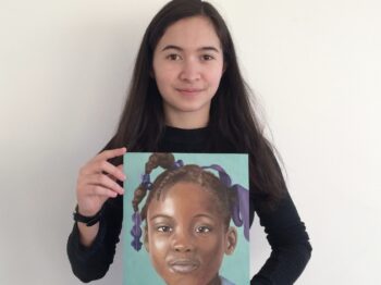 Audrey Gatta holds painting of young girl in hands while looking directly into the camera with a closed grin.
