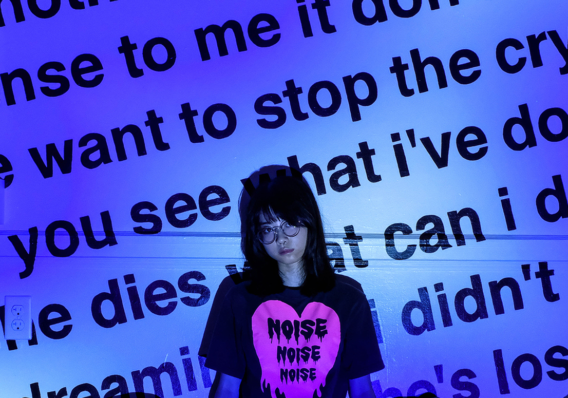 Woman wearing black in a dark room in front of a blue wall with large words on it. 