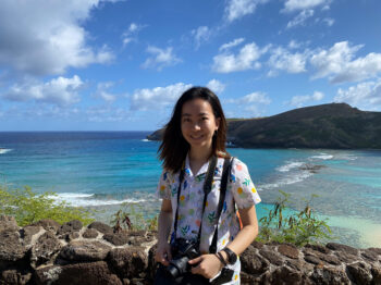 Stephanie Fu smiles at camera while holder her camera in from of the ocean.