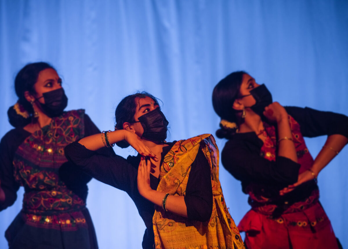 Three women in colorful clothing dancing in front of a blue background, wearing face masks and looking up and to their left.