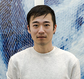 Timothy Lee in a white sweater looking into the camera in front of a blue and white background.