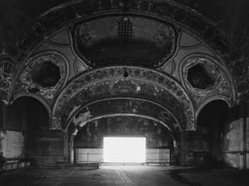 Black and white photograph of large empty theater with a decorative vaulted ceiling and a glowing white screen at the vanishing point.